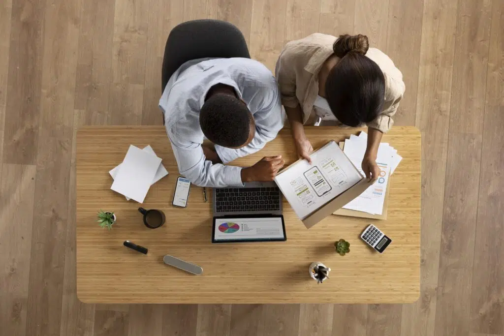 Top view of colleagues sitting next to each at a desk, looking at a laptop and documents.